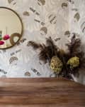 Vase of pink tulips on wooden table with round mirror and patterned wallpaper in background.
