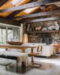 Rustic dining room with wooden table, fur-covered bench, stone fireplace, and large windows overlooking a patio.