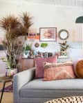 Gray sofa with pink and orange pillows, surrounded by plants, art, and shelves in a cozy living room.