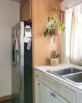 Kitchen sink with dish rack, hanging plant, stainless steel fridge, and window with light curtains.