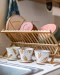 Dish rack with pink and green bowls, cups with handprint designs, and potted plants by a kitchen sink.