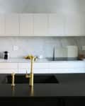 Modern kitchen with white cabinets, marble backsplash, black countertop, gold faucet, wine bottle, and glass.