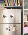 Stainless steel fridge with googly eyes, books on top, and pantry shelves with jars and a pink Smeg toaster.