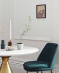 Round marble table with gold base, teal velvet chairs, vases, candle, and framed art in a bright room.