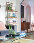 A clear, curved coffee table with books stacked on top and a tray of plants