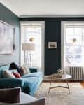Cozy living room with teal sofa, abstract art, white fireplace, and large windows with hanging plants.
