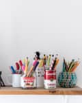 Shelf with Eiffel Tower model, framed art, and colorful pens in jars, including Nutella and Campbell's soup cans.