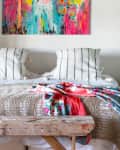 Bedroom with a colorful abstract painting, striped pillows, wooden bench, and floral quilt on the bed.