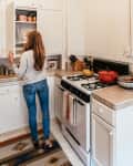 Woman in a kitchen reaching for mugs in a cabinet, with a stove, red pot, and patterned rug visible.