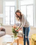 Woman arranging colorful flowers in a vase on a wooden coffee table in a bright living room with large windows.