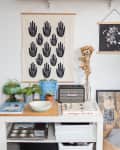 Shelf with plants, books, and art above a table with a radio, framed prints, and a tapestry of black handprints.