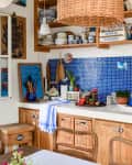 Cozy kitchen with blue mosaic backsplash, wooden cabinets, wicker baskets, and colorful decor on open shelves.