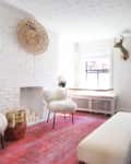 Living room with white brick fireplace, red rug, black sofa, fluffy chairs, and a mounted deer head. Kitchen visible in background.
