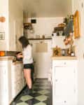 Woman cooking in a narrow kitchen with white cabinets, checkered floor, and hanging copper pots.