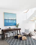 Modern living room with geometric rug, wooden desk, abstract painting, and white staircase with glass railing.