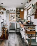 Cozy kitchen with wooden floors, open shelves, a cat on the counter, and a red backpack hanging by the door.