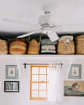 Woven baskets on a shelf above a window with framed art and books, under a ceiling fan in a bright room.