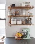 Kitchen counter with mint green mixer, fruit basket, and wooden shelves holding jars of ingredients.