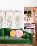 Green velvet sofa with round pink pillows, wooden table, ornate white room divider, and hanging plants.