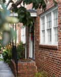 Brick house entrance with white-framed windows, surrounded by lush greenery and a small porch.