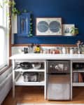 Kitchen with blue wall, white counter, small fridge, spice jars, cookware, and framed art.