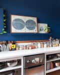 Cozy kitchen with blue walls, white shelves, vintage stove, and a small dining area with floral wallpaper and black chairs.