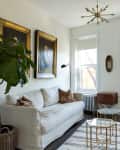 Cozy living room with white sofa, tan armchair, large plant, and framed portraits on walls, featuring a geometric coffee table.