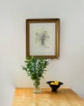 Framed floral sketch above a wooden table with a vase of greenery and a black bowl of lemons.