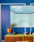 Blue wall with a pool painting above a green sideboard, featuring a bowl of oranges, a plant, and silver tea set.