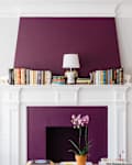 White fireplace with purple accent wall, books, lamp, and orchid, flanked by blue chairs and framed art.