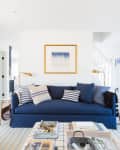 Coastal-themed living room with blue sofa, striped pillows, floral armchairs, and nautical artwork on white shiplap walls.