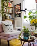 Living room with pink chair, face pillow, cat on perch, sunflowers in vase, and bookshelves near kitchen.