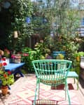 Colorful garden patio with wicker sofa, green chairs, blue bench, vibrant plants, and patterned rug under a green umbrella.