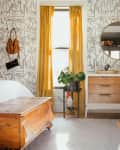 Bedroom with yellow curtains, abstract wallpaper, wooden dresser, round mirror, and wicker basket.