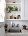 White kitchen with open shelves, potted plants, a sink, and a dish rack holding dishes and a cutting board.