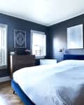 Bedroom with dark blue walls, white bedding, wooden dresser, two framed artworks, and brass wall lamps.