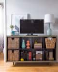TV on a shelf with wicker baskets, books, and decor; large mirror reflects a blue dresser and framed art.