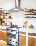 Modern kitchen with stainless steel appliances, wooden cabinets, open shelves with jars, and a bowl of fruit on the counter.