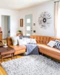 Mid-century living room with a brown leather sectional, wooden piano, geometric pillows, and a sunburst wall clock.