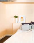 Bed with white bedding, gray and dark pillows, peach wall, abstract art, plant, and lamp on headboard shelf.