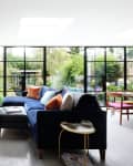 Living room with blue sectional sofa, colorful cushions, wall-mounted guitar, wooden sideboard, and large garden-view windows.
