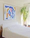 Bedroom with a white bed, abstract wall art, wooden nightstand, lamp, and large plant by a window.