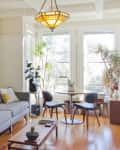 Living room with gray sofa, wooden bookshelf, round dining table, chairs, large plants, and a geometric ceiling light.