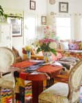Colorful living room with floral sofa, patterned cushions, vibrant rugs, and a table with a vase of flowers.