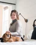 Woman sitting on a bed with a small brown dog, white shiplap walls, and a black wall lamp.