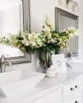 Bathroom with dual sinks, large mirror, white flowers in a vase, and shelves with decor above the toilet.