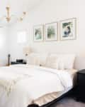 Bright bedroom with a large bed, black nightstands, beige pillows, a chandelier, and framed photos on the wall.