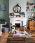 Living room with a white fireplace, round mirror, bookshelves, and a wooden coffee table with books and plants.