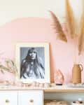Shelf with framed black-and-white portrait, pampas grass in vase, and decorative items against a pink semicircle wall.