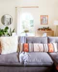 Gray sectional sofa with colorful pillows, plants, and wall art in a bright living room with arched window and wooden floor.